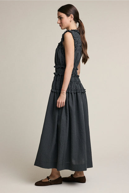 A women's sleeveless maxi dress in a dark gray and black plaid pattern featuring a high ruffled neckline, a fully elasticized smocked bodice, and a flowing tiered skirt with delicate ruffle trim.
