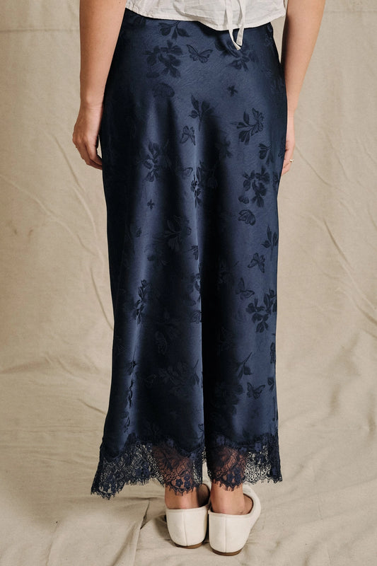 A women's navy blue satin bias-cut slip midi skirt featuring an elastic waistband and delicate scalloped lace trim along the hemline.