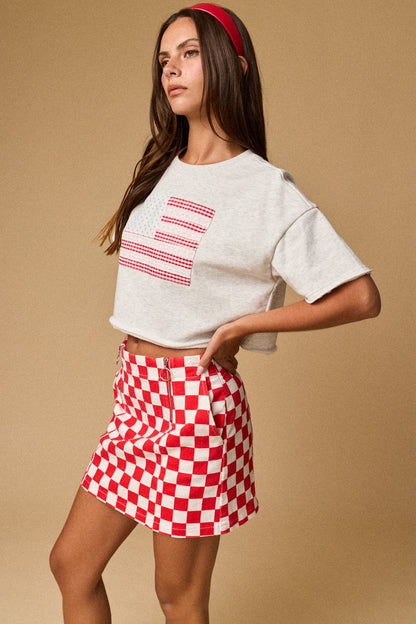 A women's light gray short-sleeve graphic t-shirt featuring a relaxed fit, a raw cut cropped hemline, and a textured patchwork American flag design made from blue star print, red gingham, and white eyelet lace fabrics.