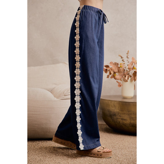 A women's medium wash wide-leg denim pants featuring a gathered elastic drawstring waistband and white crochet daisy floral lace side seam trim.