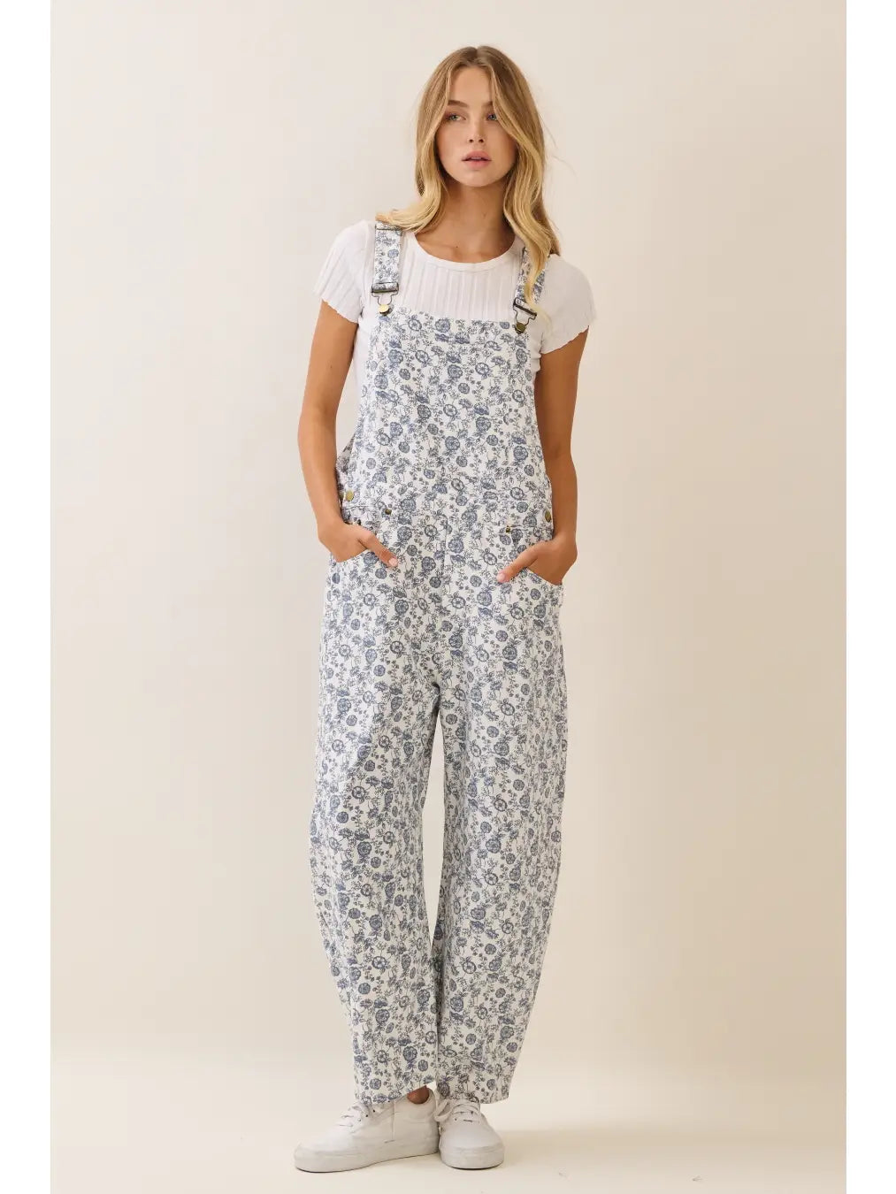 A women's sleeveless floral printed cropped overall jumpsuit in a blue and cream pattern featuring adjustable buckle shoulder straps, side button closures, utility slash pockets, and a relaxed wide-leg silhouette worn over a white short-sleeve tee.