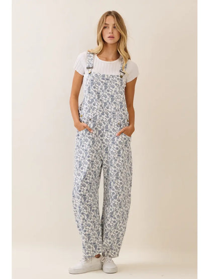 A women's sleeveless floral printed cropped overall jumpsuit in a blue and cream pattern featuring adjustable buckle shoulder straps, side button closures, utility slash pockets, and a relaxed wide-leg silhouette worn over a white short-sleeve tee.
