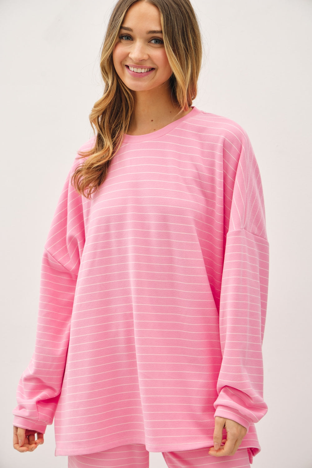 A women's relaxed long-sleeve crewneck top in bubblegum pink with ivory horizontal micro-stripes, dropped shoulders, and subtle side hem vents.