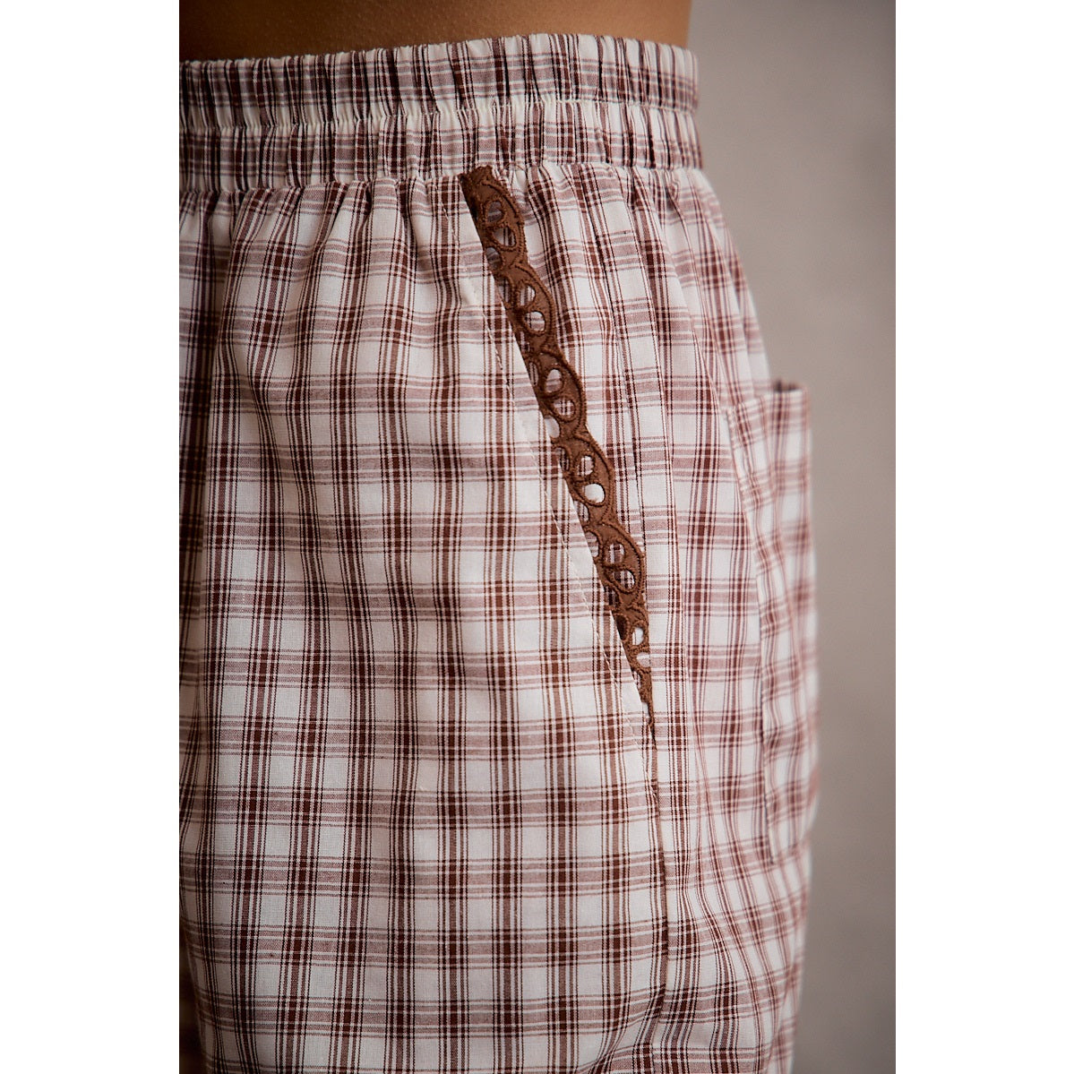 A pair of women's relaxed wide-leg brown and cream plaid pull-on pants featuring an elastic drawstring waist, side pockets, and an embroidered brown eyelet lace scalloped hem.