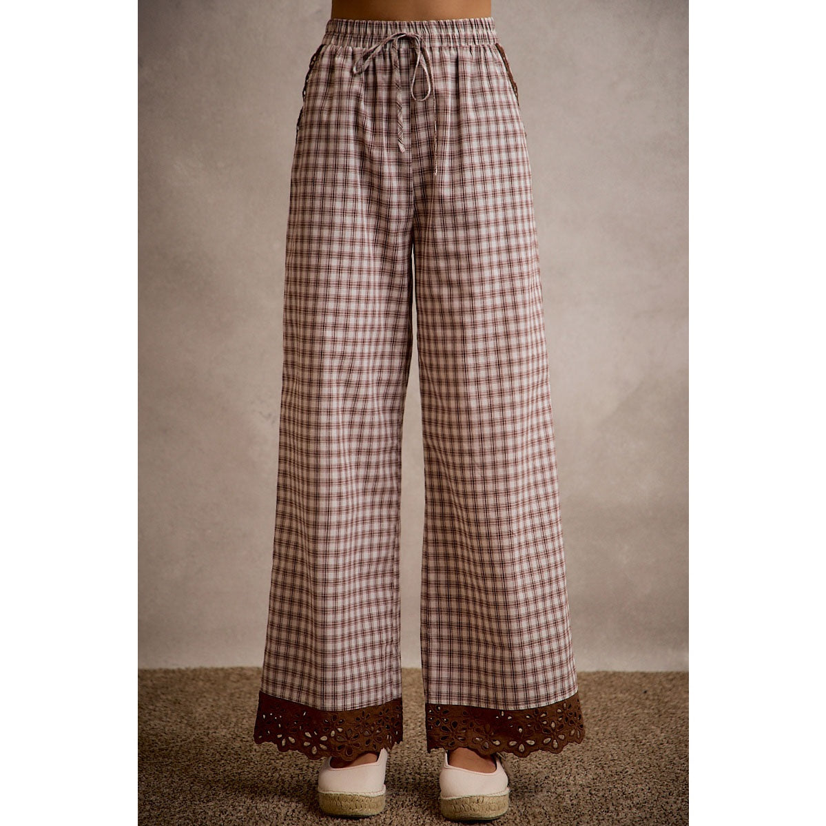 A pair of women's relaxed wide-leg brown and cream plaid pull-on pants featuring an elastic drawstring waist, side pockets, and an embroidered brown eyelet lace scalloped hem.