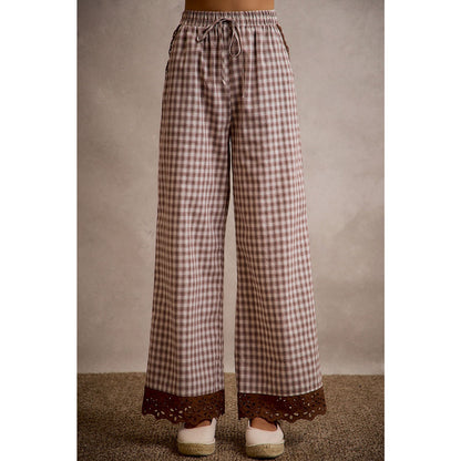 A pair of women's relaxed wide-leg brown and cream plaid pull-on pants featuring an elastic drawstring waist, side pockets, and an embroidered brown eyelet lace scalloped hem.