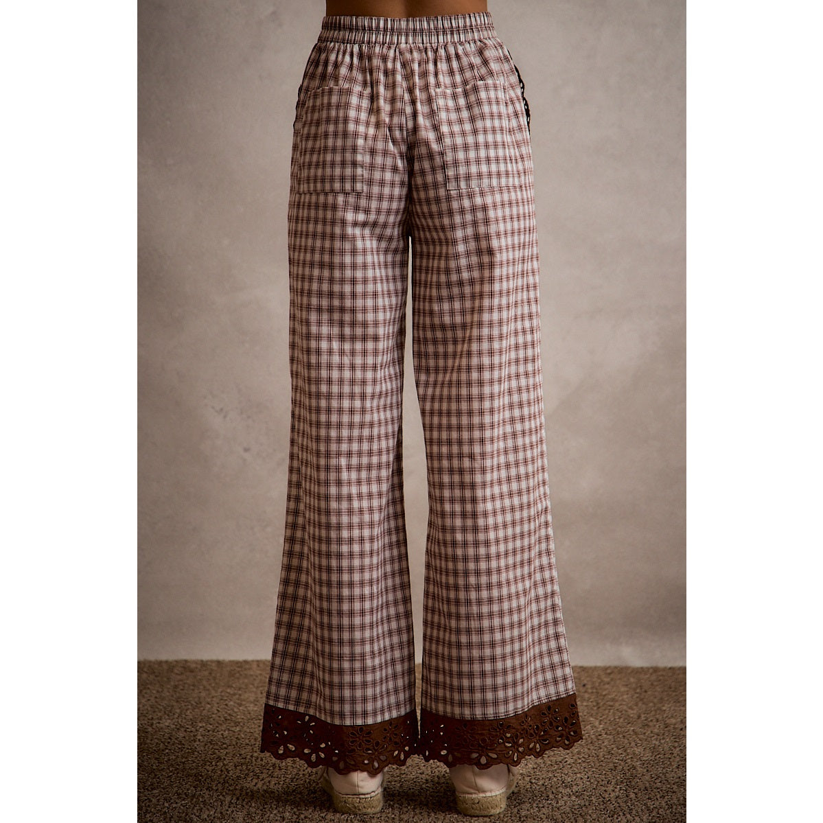A pair of women's relaxed wide-leg brown and cream plaid pull-on pants featuring an elastic drawstring waist, side pockets, and an embroidered brown eyelet lace scalloped hem.