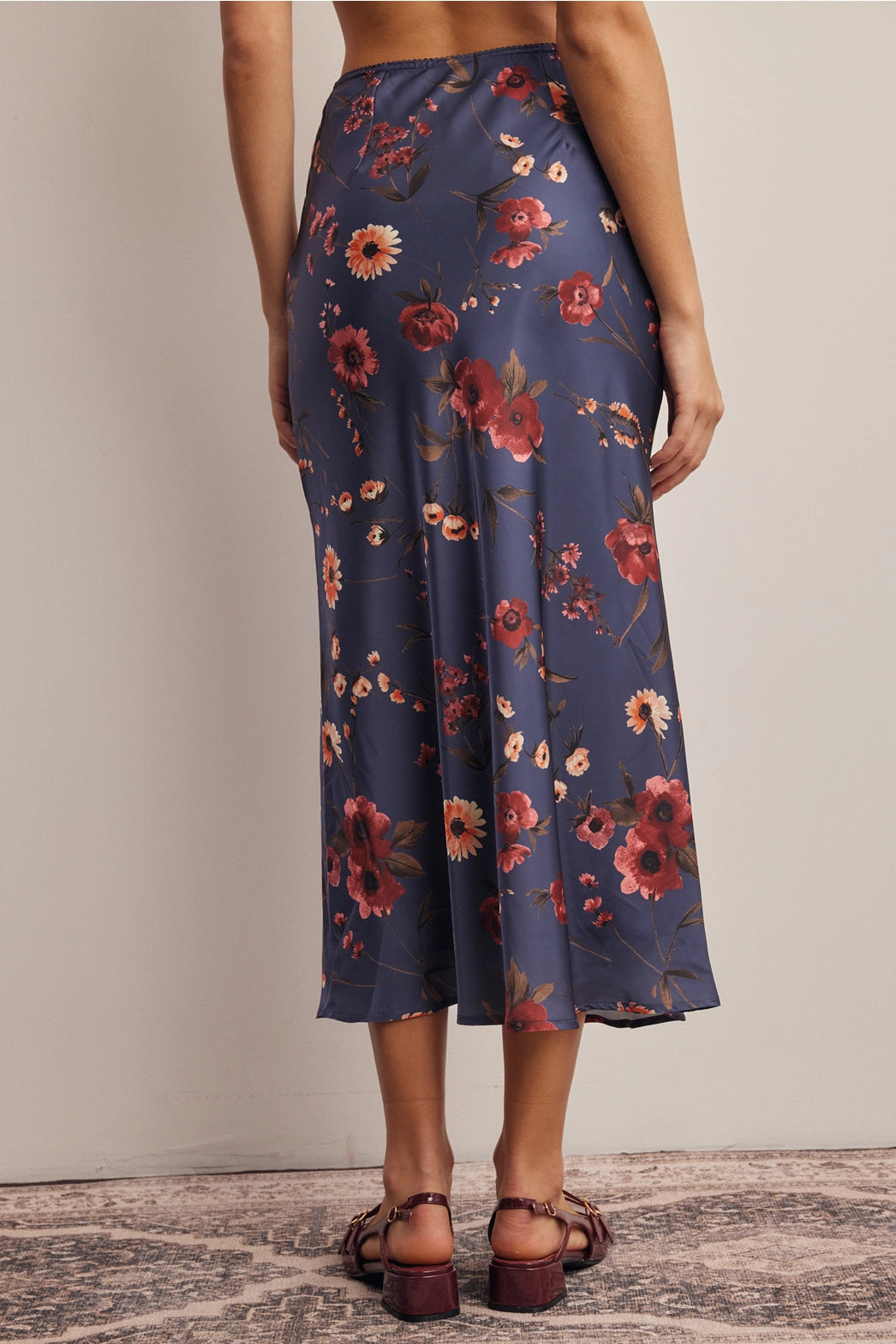 A women's bias-cut satin midi skirt in a midnight navy blue floral print featuring warm rust and amber blossoms with an elasticized waistband.