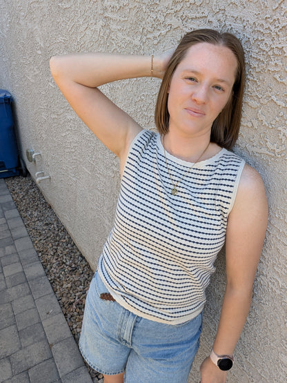 A sleeveless horizontal stripe knit sweater tank featuring a high boatneck, delicate ribbed trim at the hem and armholes, and a neutral oatmeal and navy color palette.