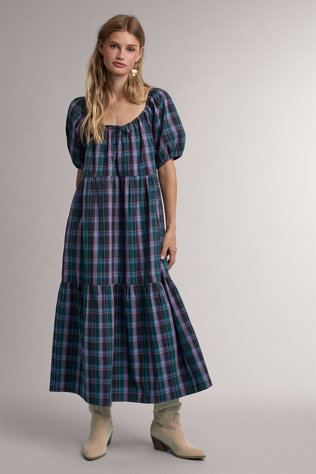 A women's tiered midi-length dress in a green and violet plaid pattern featuring short puff sleeves, a gathered scoop neck with a tie detail, and functional side pockets.