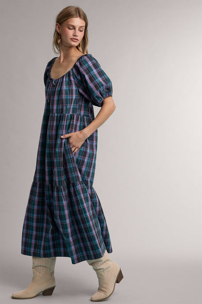 A women's tiered midi-length dress in a green and violet plaid pattern featuring short puff sleeves, a gathered scoop neck with a tie detail, and functional side pockets.