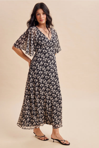 A women's v-neck maxi dress in a black and cream floral print featuring short flutter sleeves and an empire waist silhouette.