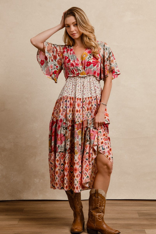 A short-sleeve tiered patchwork midi dress in a rose pink floral and geometric print featuring a deep V-neckline, flutter sleeves, and an elastic waist.