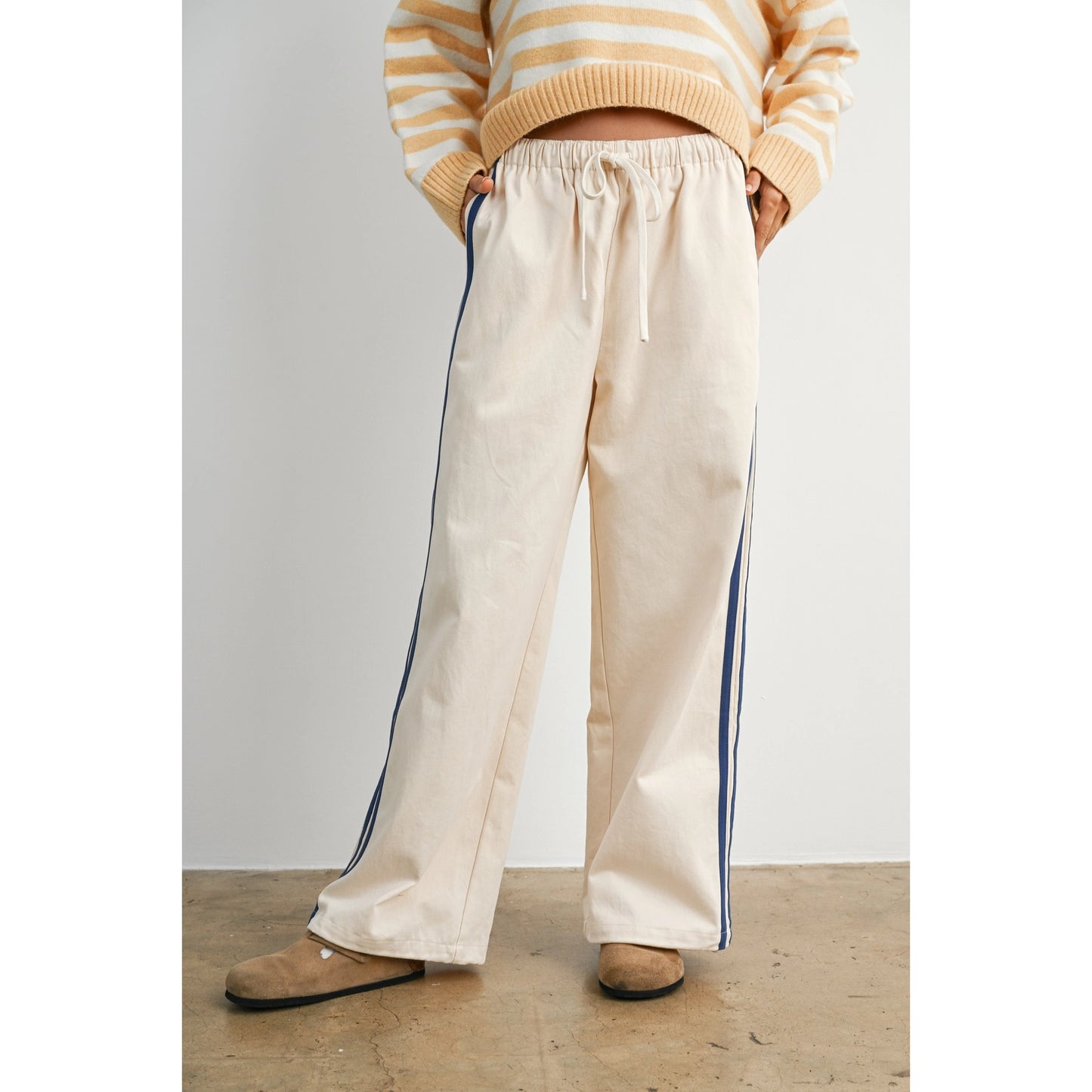 A pair of women's wide-leg relaxed cream track pants featuring double navy blue side stripes, an elastic drawstring waistband, and a mid-rise fit.