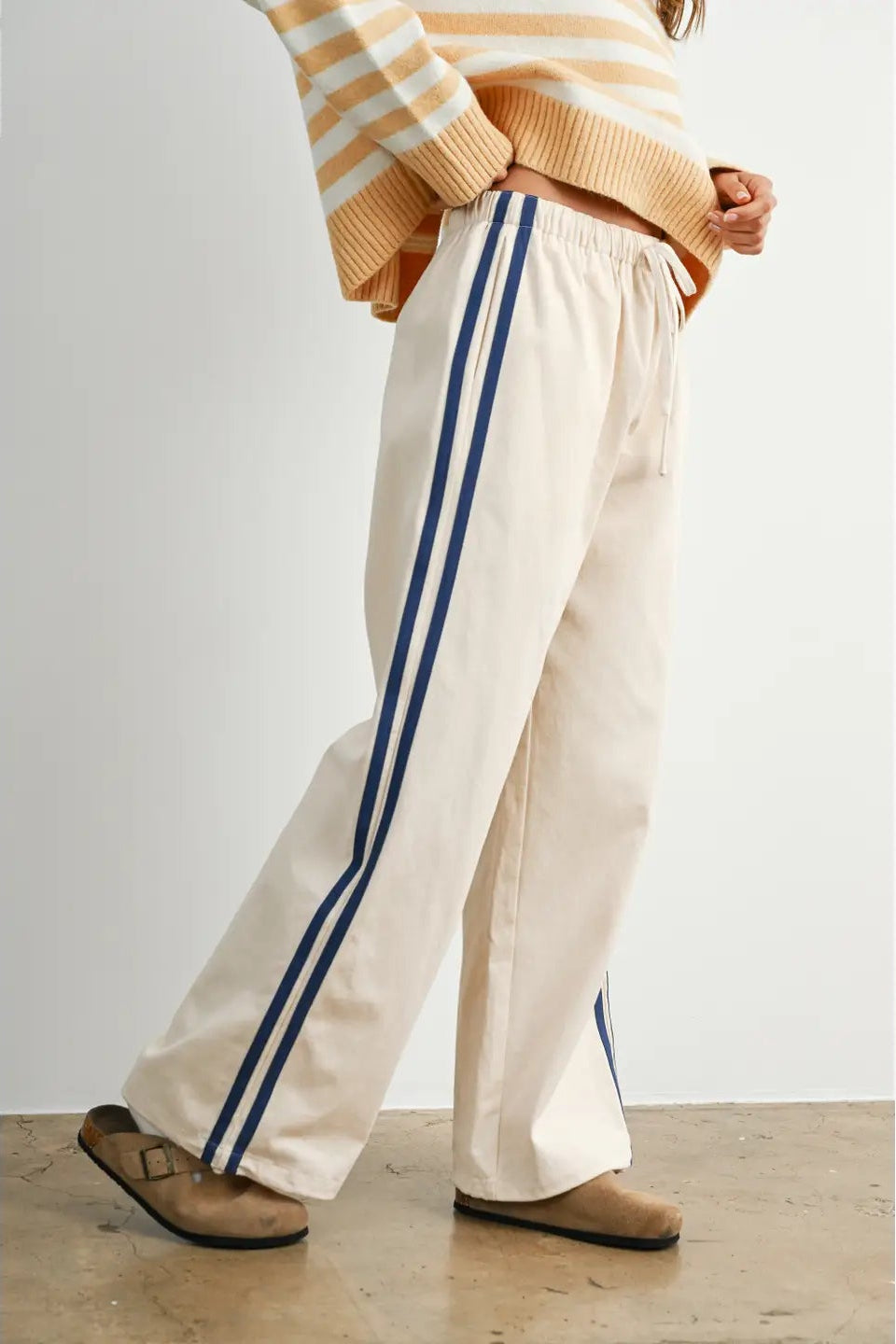 A pair of women's wide-leg relaxed cream track pants featuring double navy blue side stripes, an elastic drawstring waistband, and a mid-rise fit.