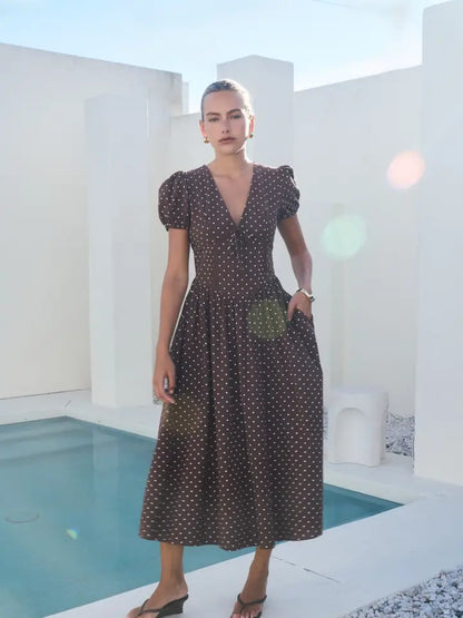 A short-sleeve brown and white polka dot midi dress featuring a V-neckline with a keyhole front tie detail, structured puff sleeves, and functional side pockets.