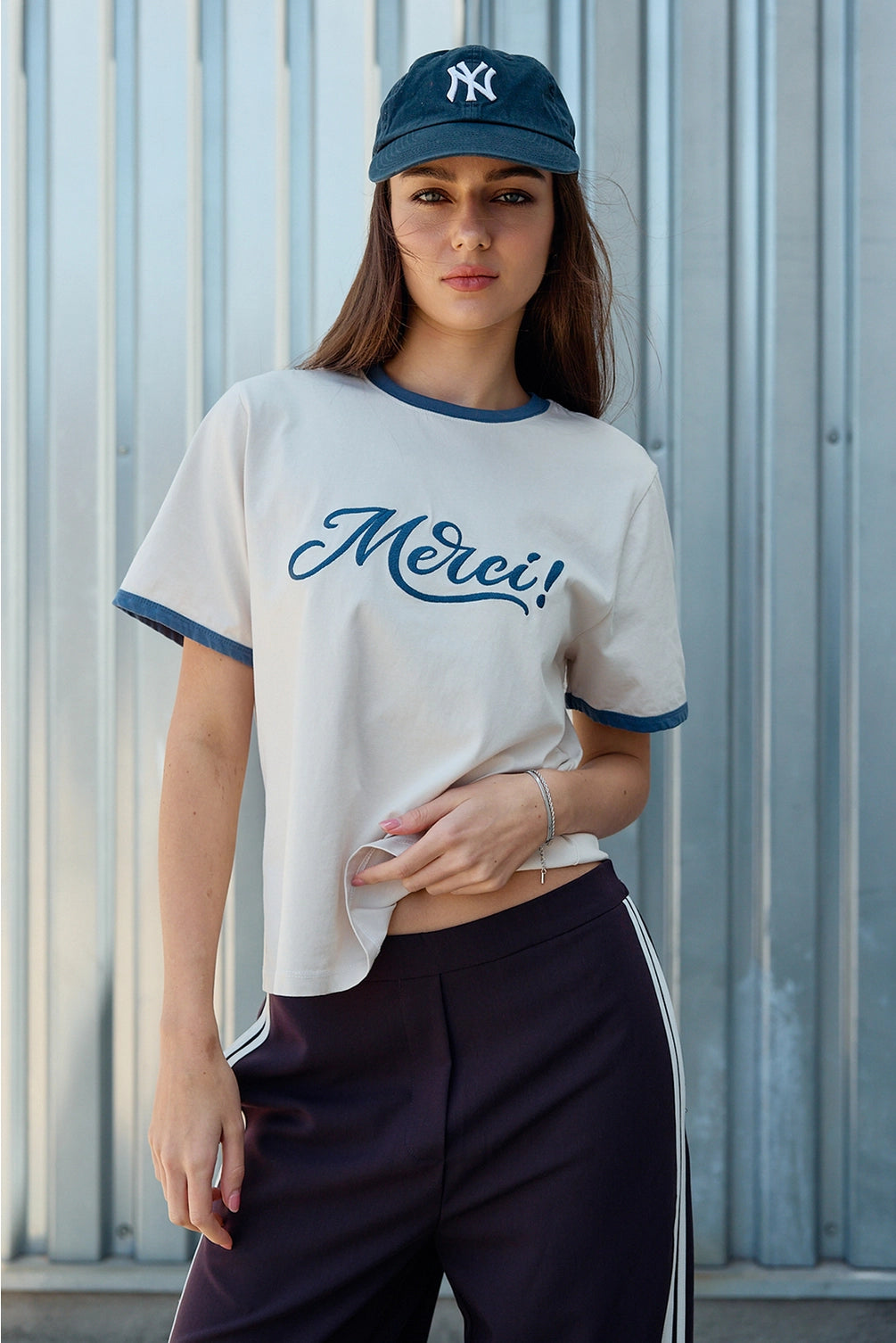 A women's light sand crewneck ringer t-shirt featuring deep blue contrast trim along the neckline and sleeves, short sleeves, and cursive blue embroidered script across the chest that says, Merci. 
