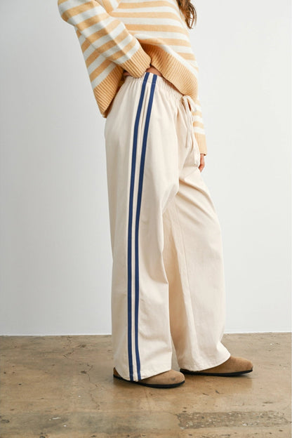 A pair of women's wide-leg relaxed cream track pants featuring double navy blue side stripes, an elastic drawstring waistband, and a mid-rise fit.