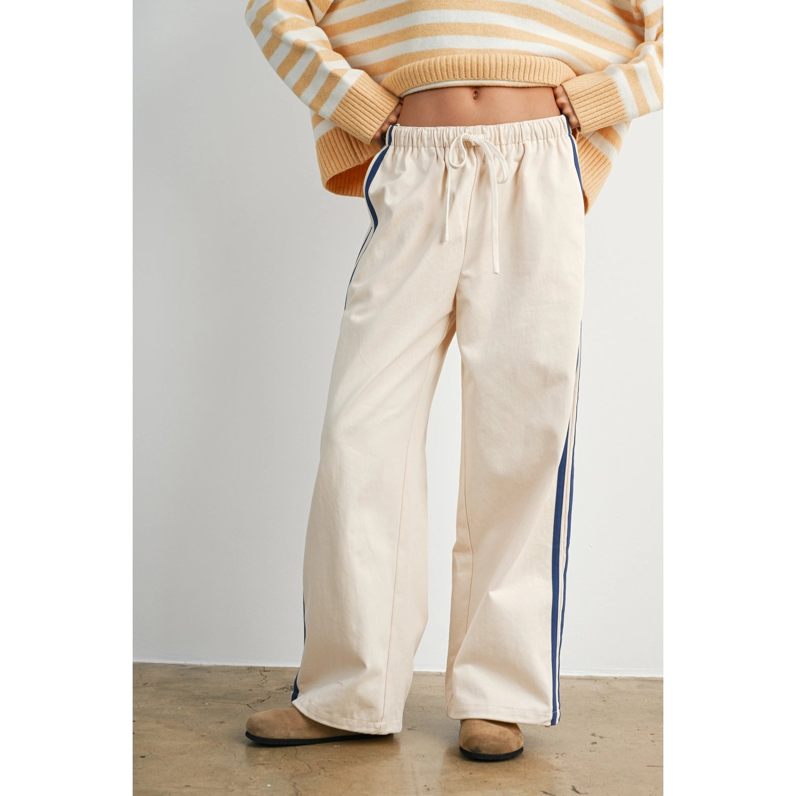 A pair of women's wide-leg relaxed cream track pants featuring double navy blue side stripes, an elastic drawstring waistband, and a mid-rise fit.