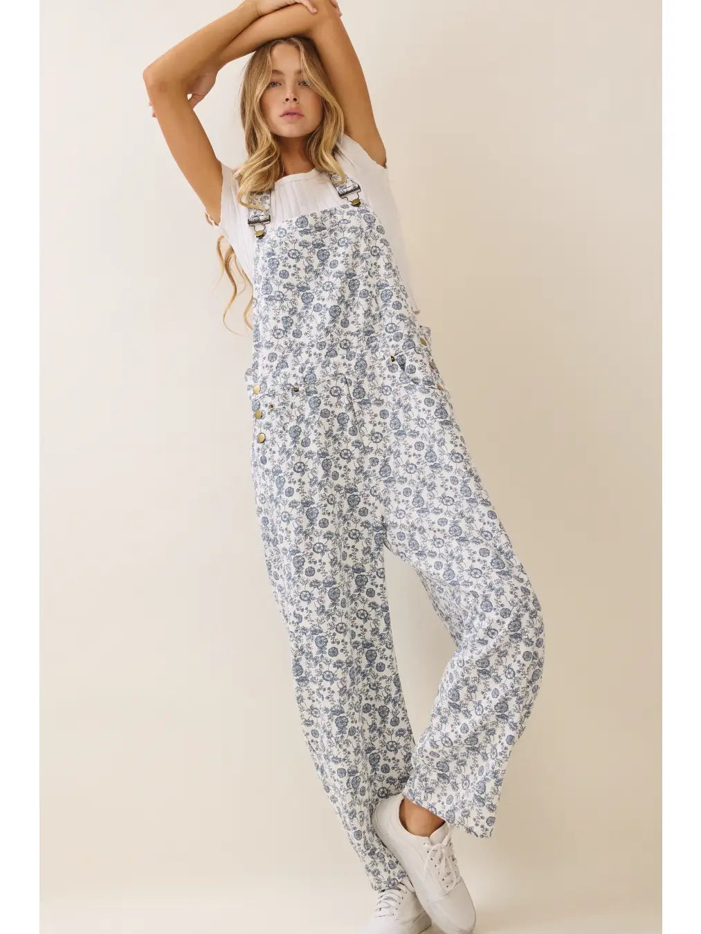 A women's sleeveless floral printed cropped overall jumpsuit in a blue and cream pattern featuring adjustable buckle shoulder straps, side button closures, utility slash pockets, and a relaxed wide-leg silhouette worn over a white short-sleeve tee.