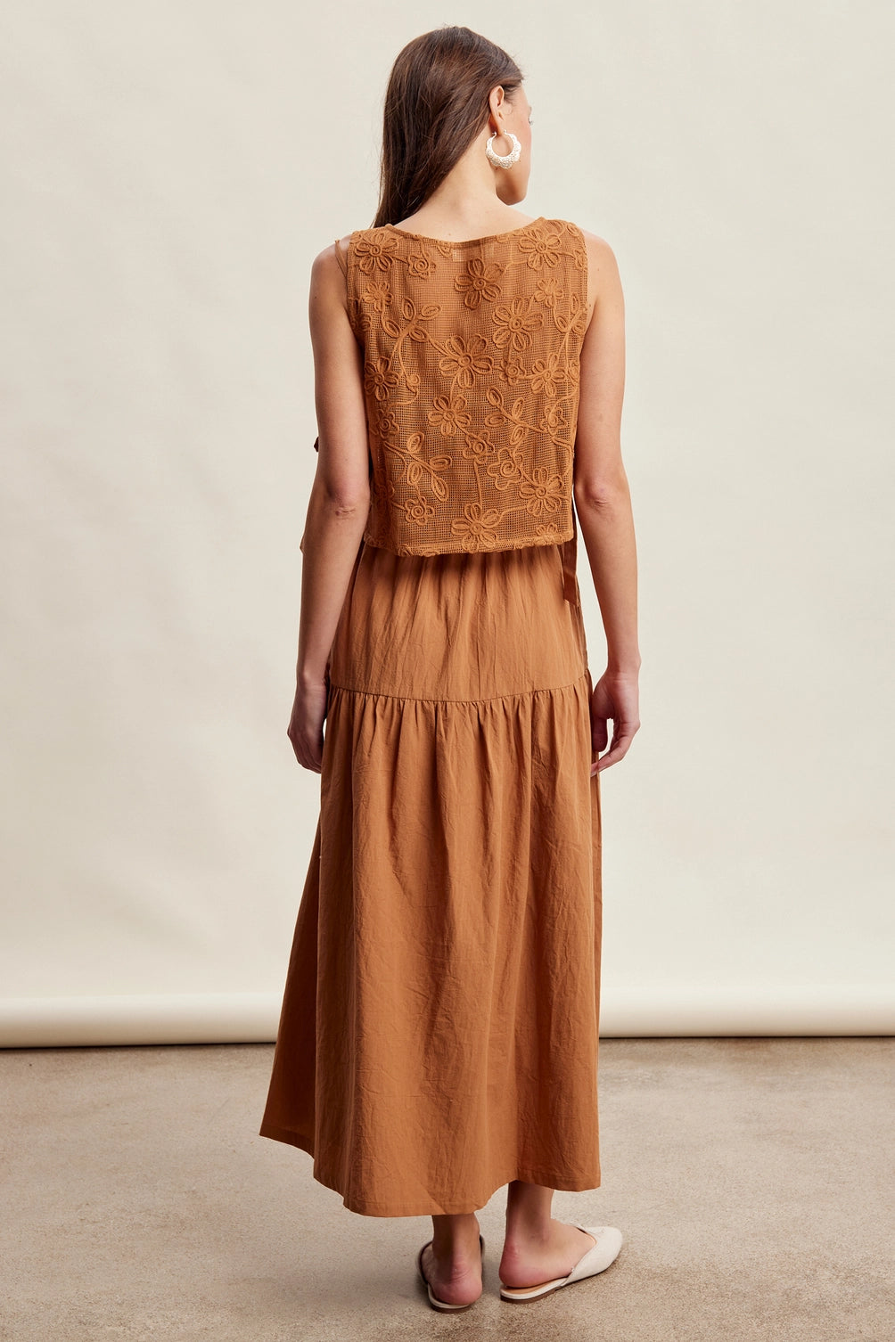 A women's brown sleeveless tiered maxi dress featuring a tonal embroidered floral lace vest overlay with adjustable side tie details.