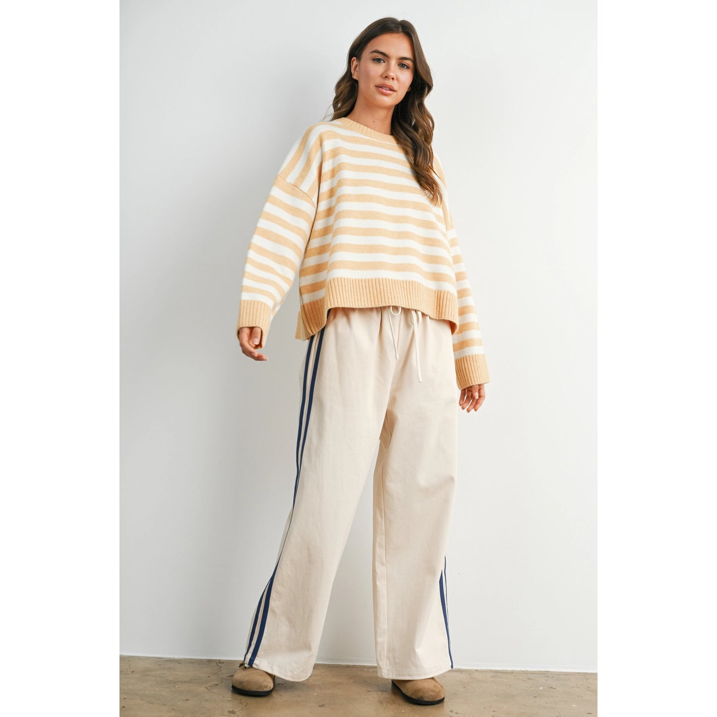 A pair of women's wide-leg relaxed cream track pants featuring double navy blue side stripes, an elastic drawstring waistband, and a mid-rise fit.