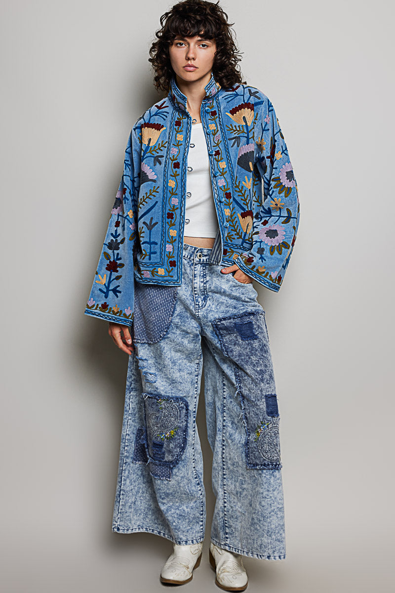 A women's dusty denim blue long-sleeve velvet jacket with allover folk floral embroidery in rust, yellow, and lavender, styled with a mandarin collar, wide cuffs, and front loop buttons.