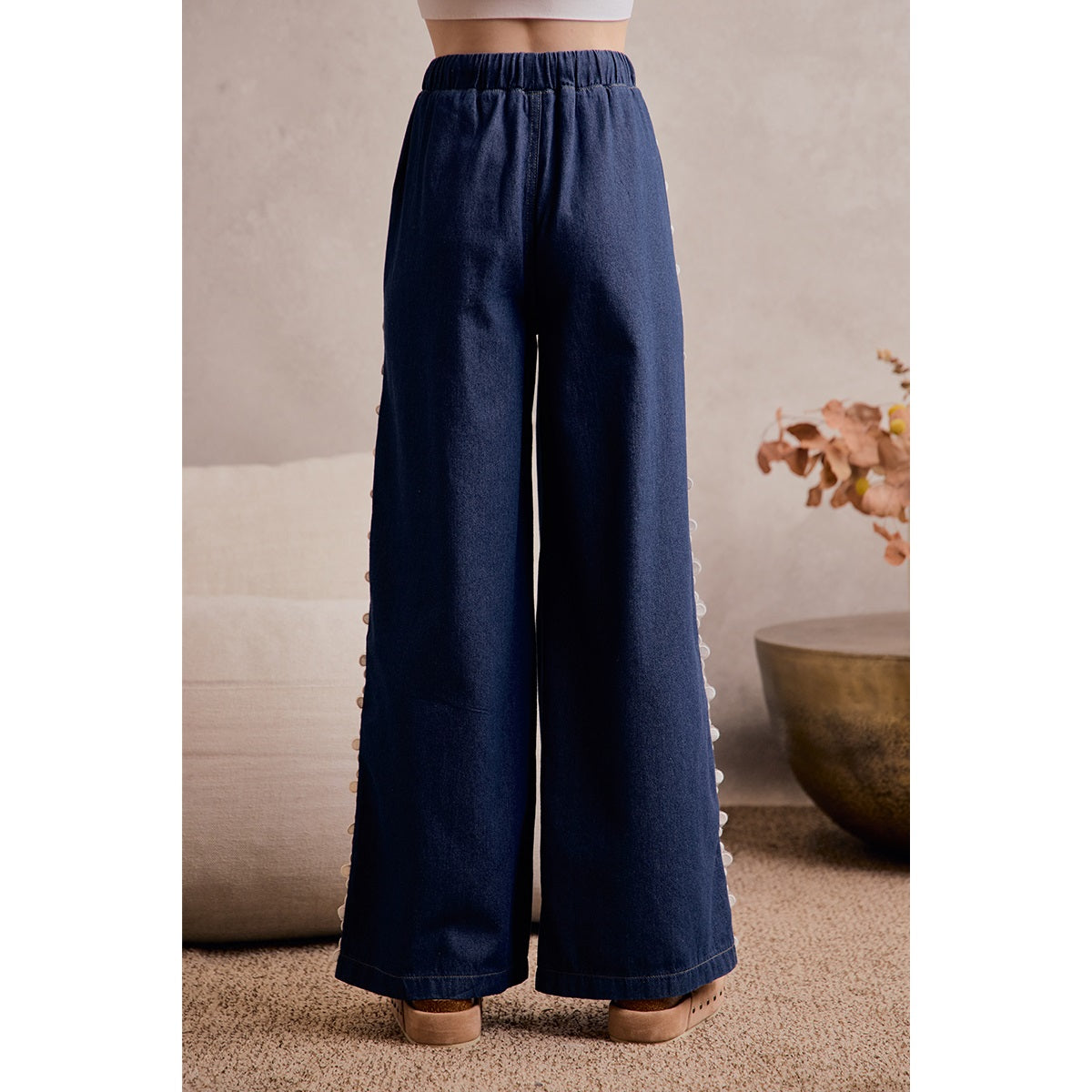 A women's medium wash wide-leg denim pants featuring a gathered elastic drawstring waistband and white crochet daisy floral lace side seam trim.