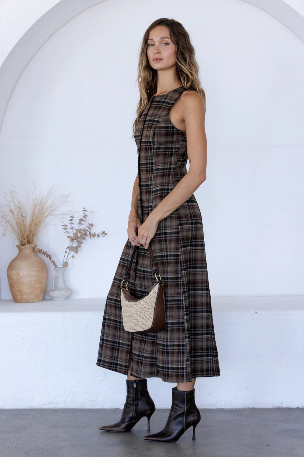 A women's sleeveless midi dress in brown and black plaid featuring a boat neckline, vertical front buttons, and a straight column silhouette.