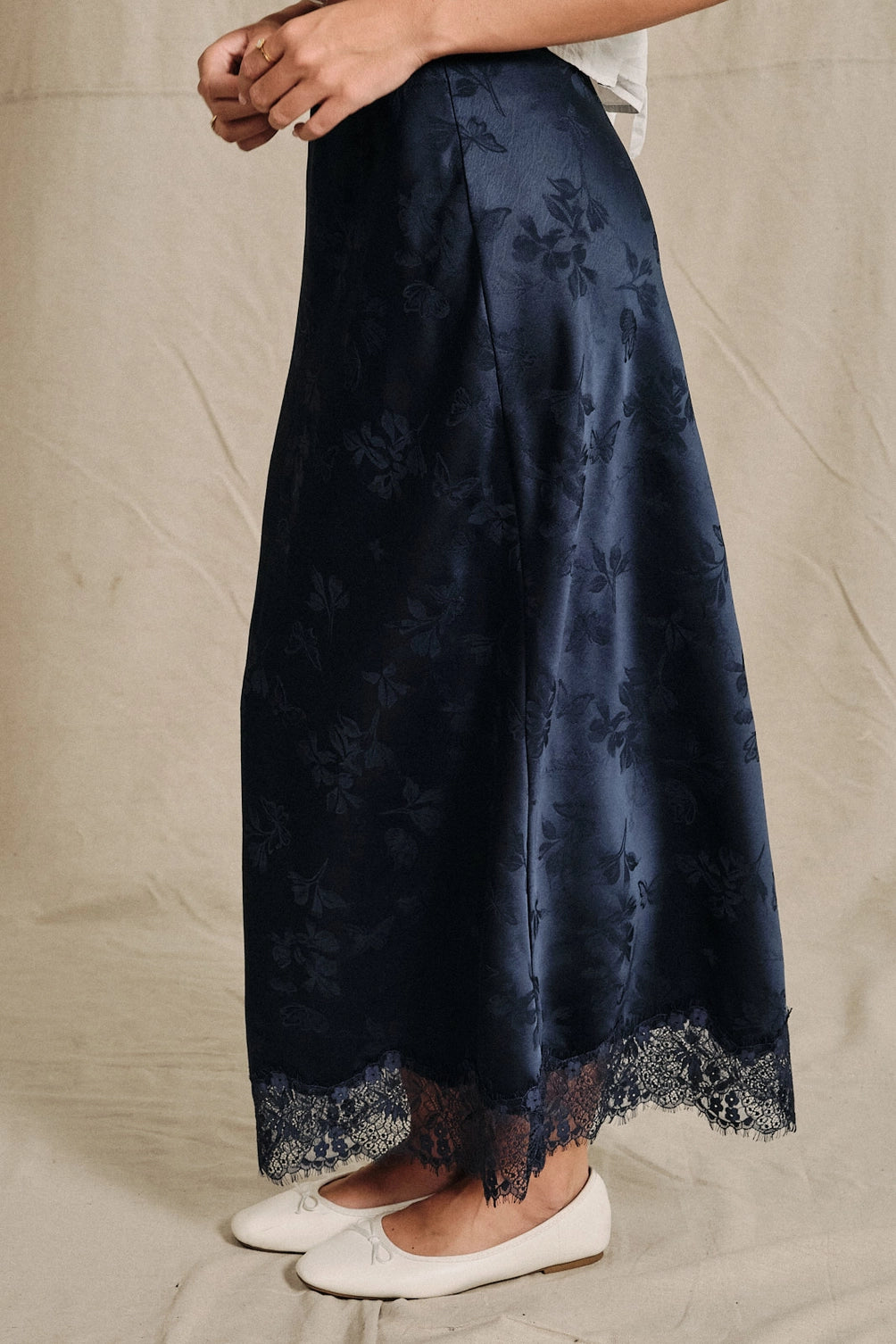 A women's navy blue satin bias-cut slip midi skirt featuring an elastic waistband and delicate scalloped lace trim along the hemline.