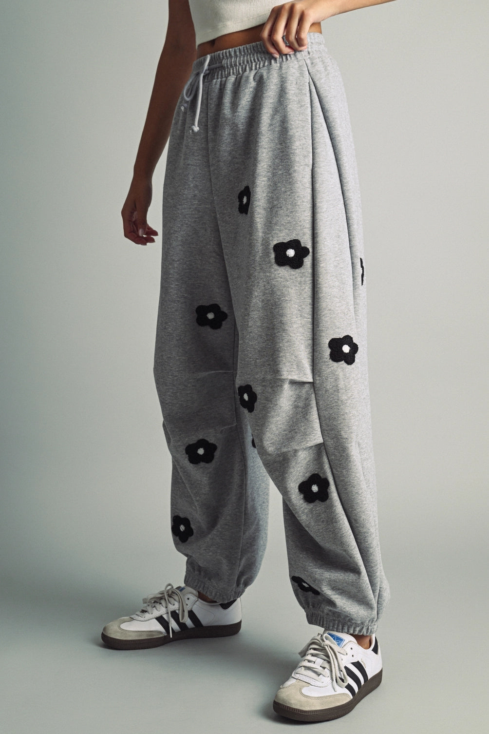 A women's heather gray wide-leg fleece sweatpants featuring an elastic drawstring waistband, relaxed barrel leg, and black chenille flower patches across the front and back.