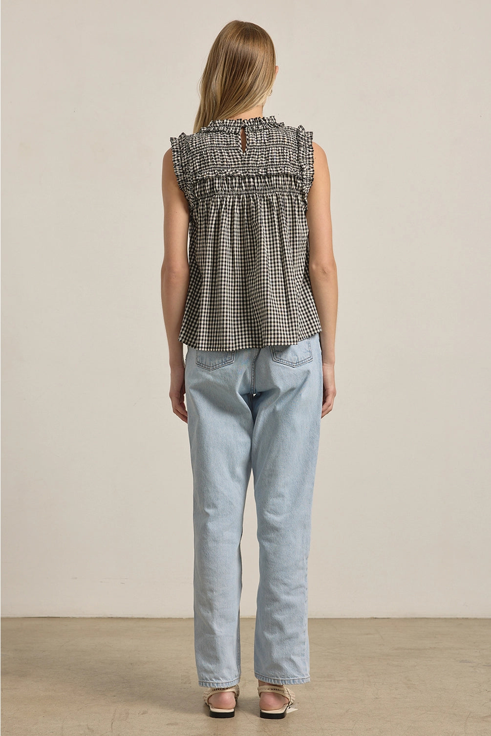 A women's sleeveless black and white gingham check blouse featuring a smocked high mock neckline, ruffle shoulder details, and a relaxed baby-doll silhouette.