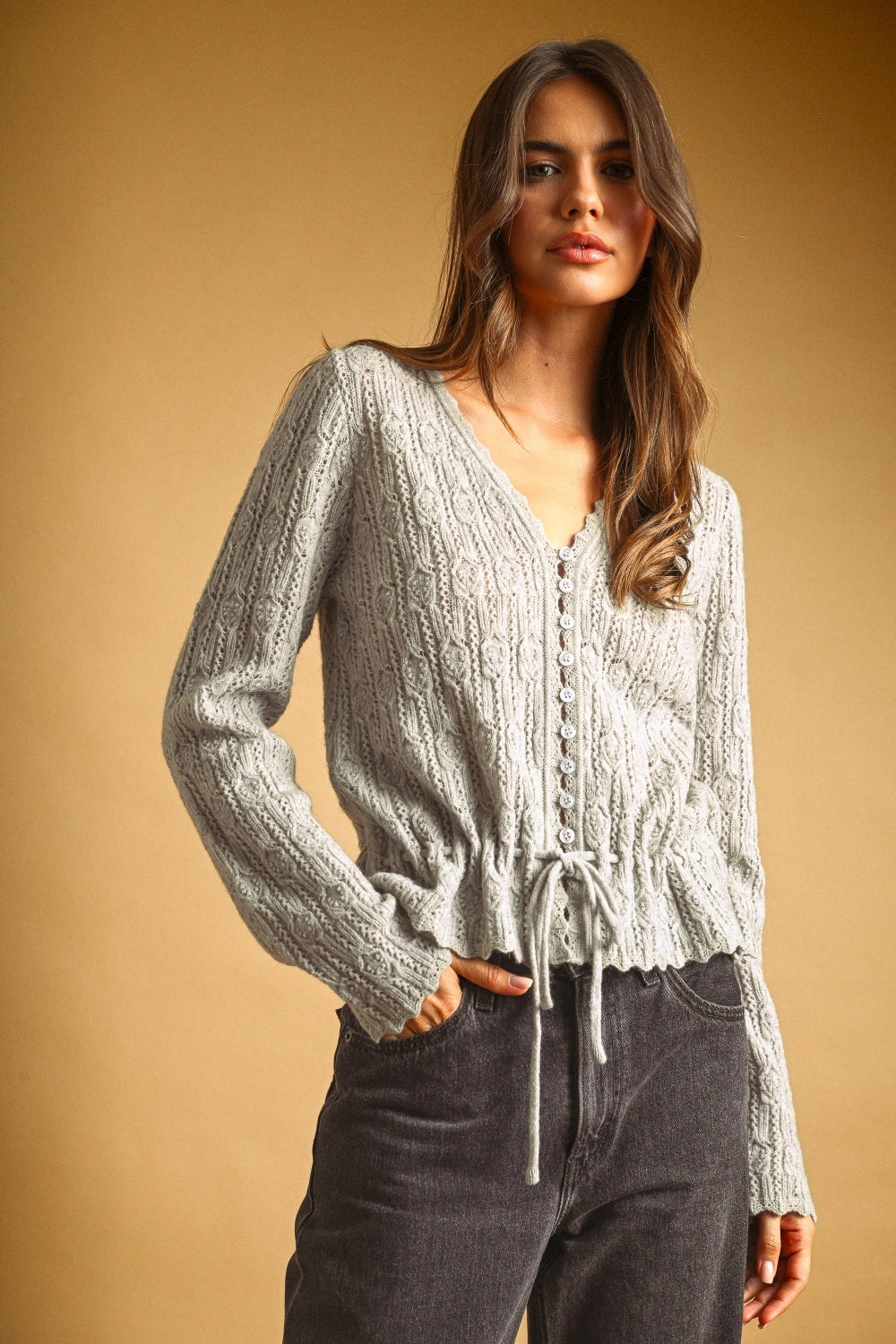 A women's light gray long-sleeve textured lace knit cardigan sweater featuring a button-down front, scalloped V-neckline, and an adjustable drawstring tie waistline.