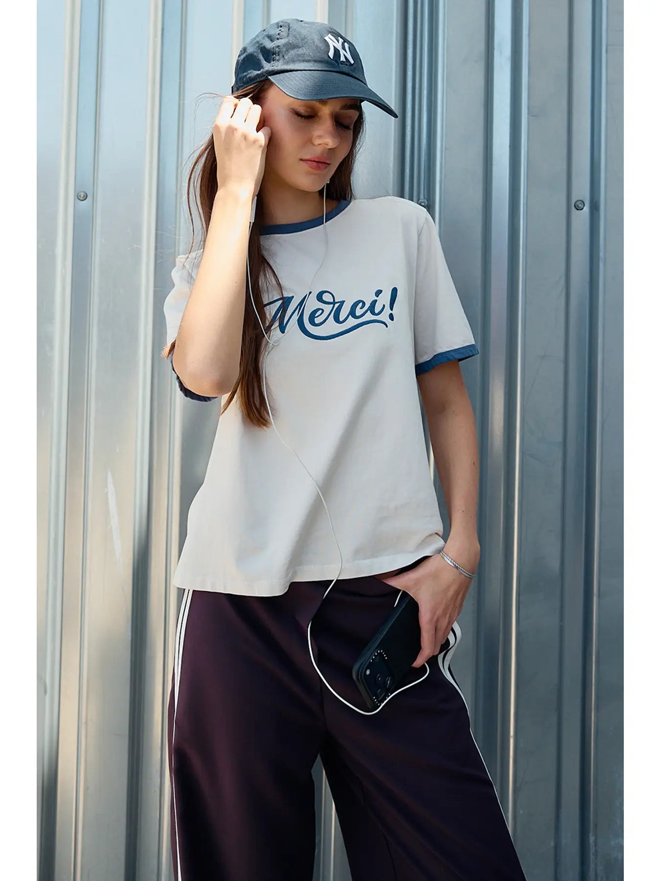 A women's light sand crewneck ringer t-shirt featuring deep blue contrast trim along the neckline and sleeves, short sleeves, and cursive blue embroidered script across the chest that says, Merci. 