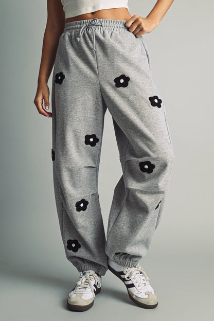 A women's heather gray wide-leg fleece sweatpants featuring an elastic drawstring waistband, relaxed barrel leg, and black chenille flower patches across the front and back.