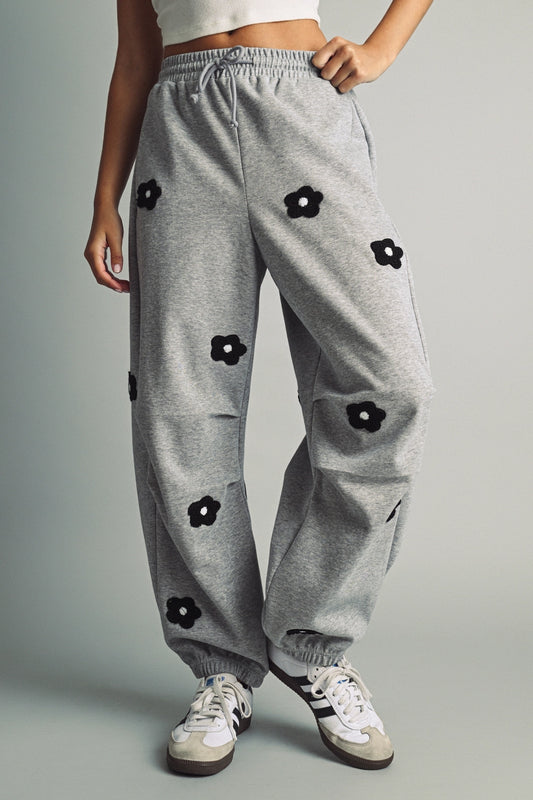 A women's heather gray wide-leg fleece sweatpants featuring an elastic drawstring waistband, relaxed barrel leg, and black chenille flower patches across the front and back.