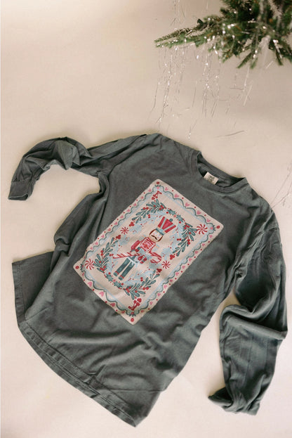 A women's gray sage green long-sleeve cotton graphic tee featuring a vintage-inspired holiday nutcracker playing card design with botanical greenery and pink accents.
