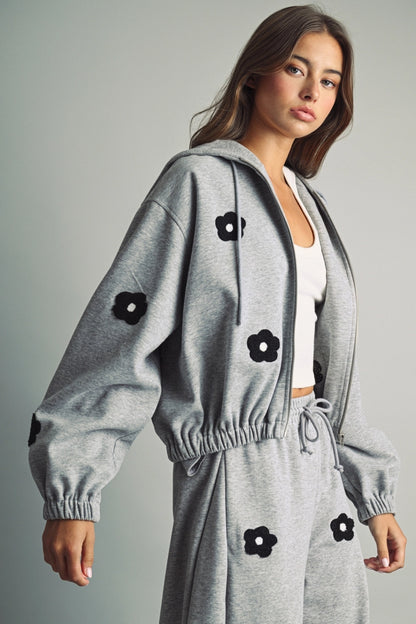 A women's heather gray full-zip hooded fleece jacket featuring dropped shoulders, ribbed hem cuffs, and black chenille flower patches on the front and sleeves.