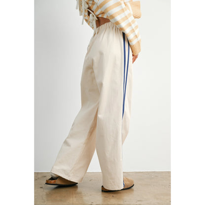 A pair of women's wide-leg relaxed cream track pants featuring double navy blue side stripes, an elastic drawstring waistband, and a mid-rise fit.