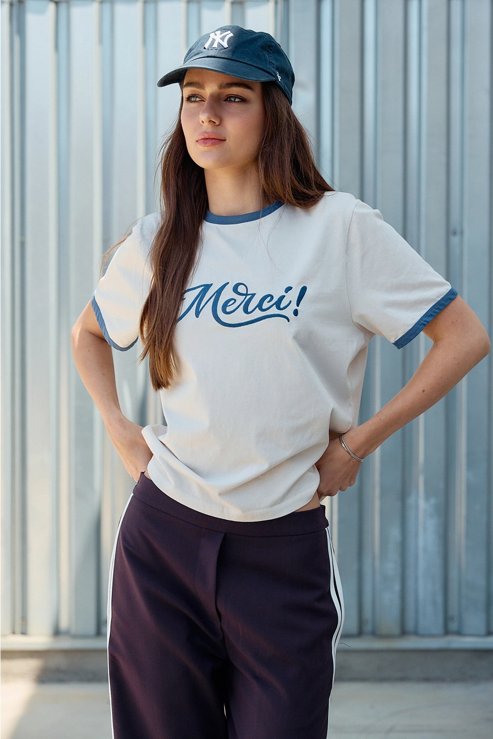 A women's light sand crewneck ringer t-shirt featuring deep blue contrast trim along the neckline and sleeves, short sleeves, and cursive blue embroidered script across the chest that says, Merci. 