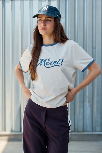 A women's light sand crewneck ringer t-shirt featuring deep blue contrast trim along the neckline and sleeves, short sleeves, and cursive blue embroidered script across the chest that says, Merci. 