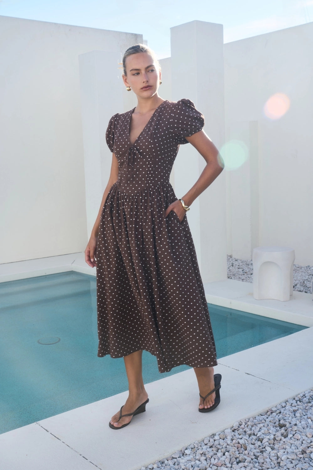 A short-sleeve brown and white polka dot midi dress featuring a V-neckline with a keyhole front tie detail, structured puff sleeves, and functional side pockets.