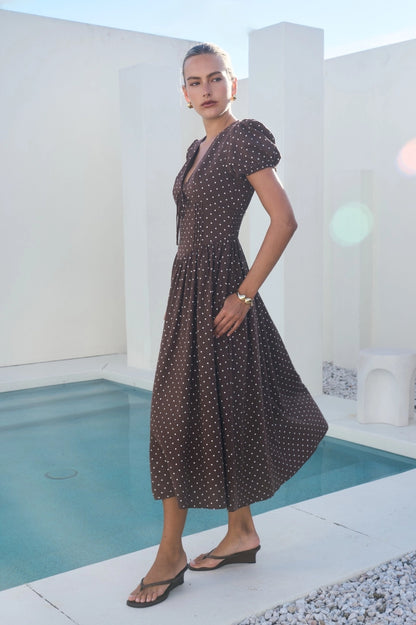 A short-sleeve brown and white polka dot midi dress featuring a V-neckline with a keyhole front tie detail, structured puff sleeves, and functional side pockets.