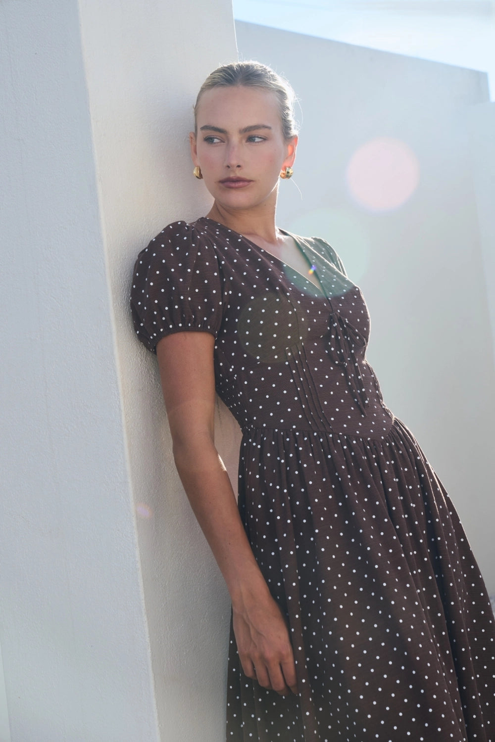 A short-sleeve brown and white polka dot midi dress featuring a V-neckline with a keyhole front tie detail, structured puff sleeves, and functional side pockets.