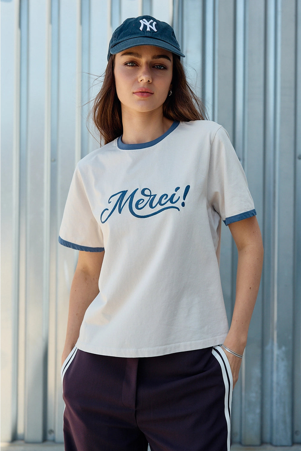 A women's light sand crewneck ringer t-shirt featuring deep blue contrast trim along the neckline and sleeves, short sleeves, and cursive blue embroidered script across the chest that says, Merci. 