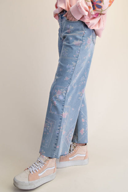 A pair of women's high-waisted wide-leg stretch denim jeans in a light wash featuring an all-over purple and pink floral print and frayed raw hems.