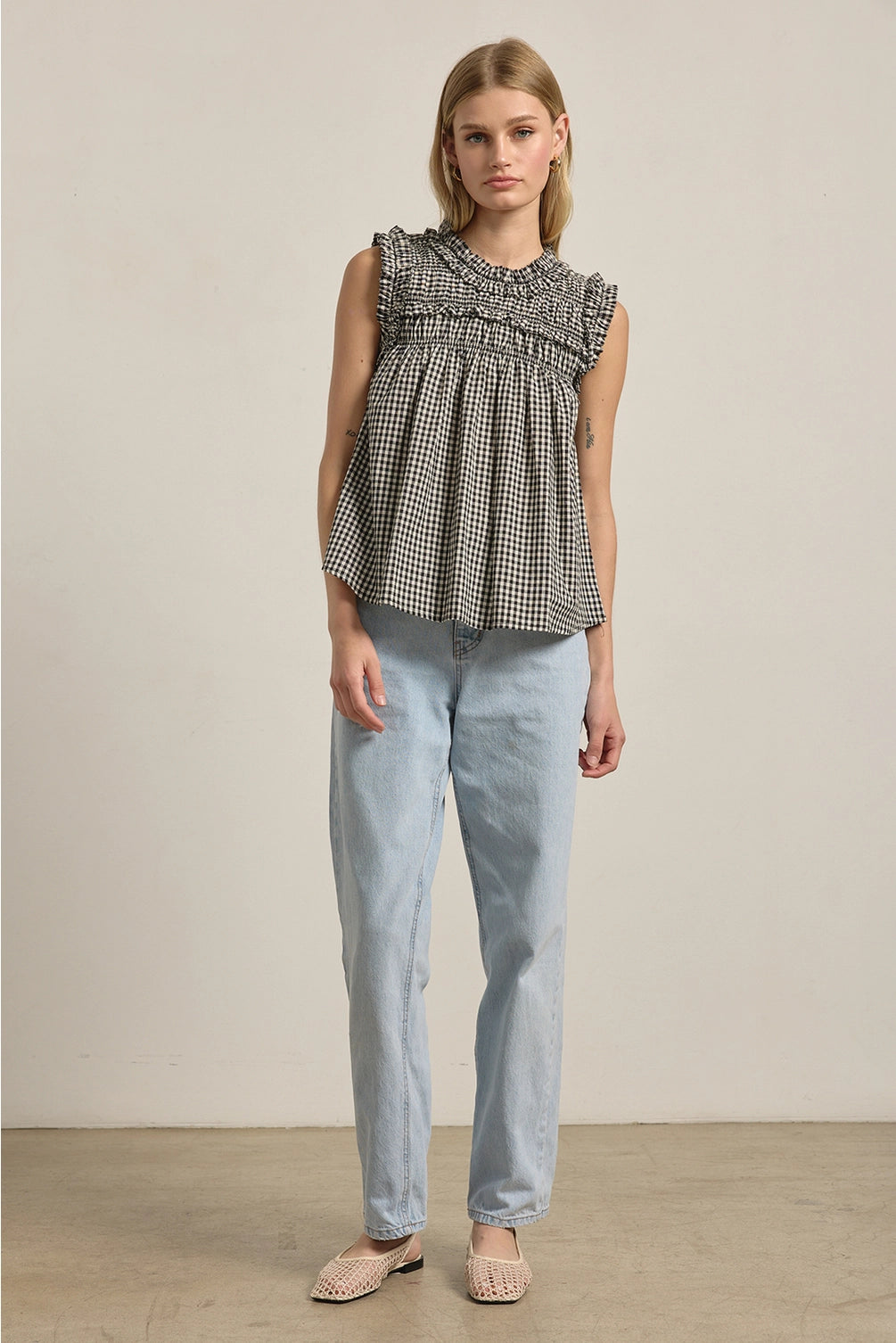 A women's sleeveless black and white gingham check blouse featuring a smocked high mock neckline, ruffle shoulder details, and a relaxed baby-doll silhouette.