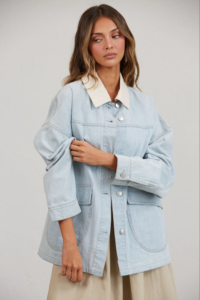 A women's relaxed oversized light wash denim utility jacket featuring a cream-colored pointed contrast collar, a button-front closure, and large front patch pockets with flaps.