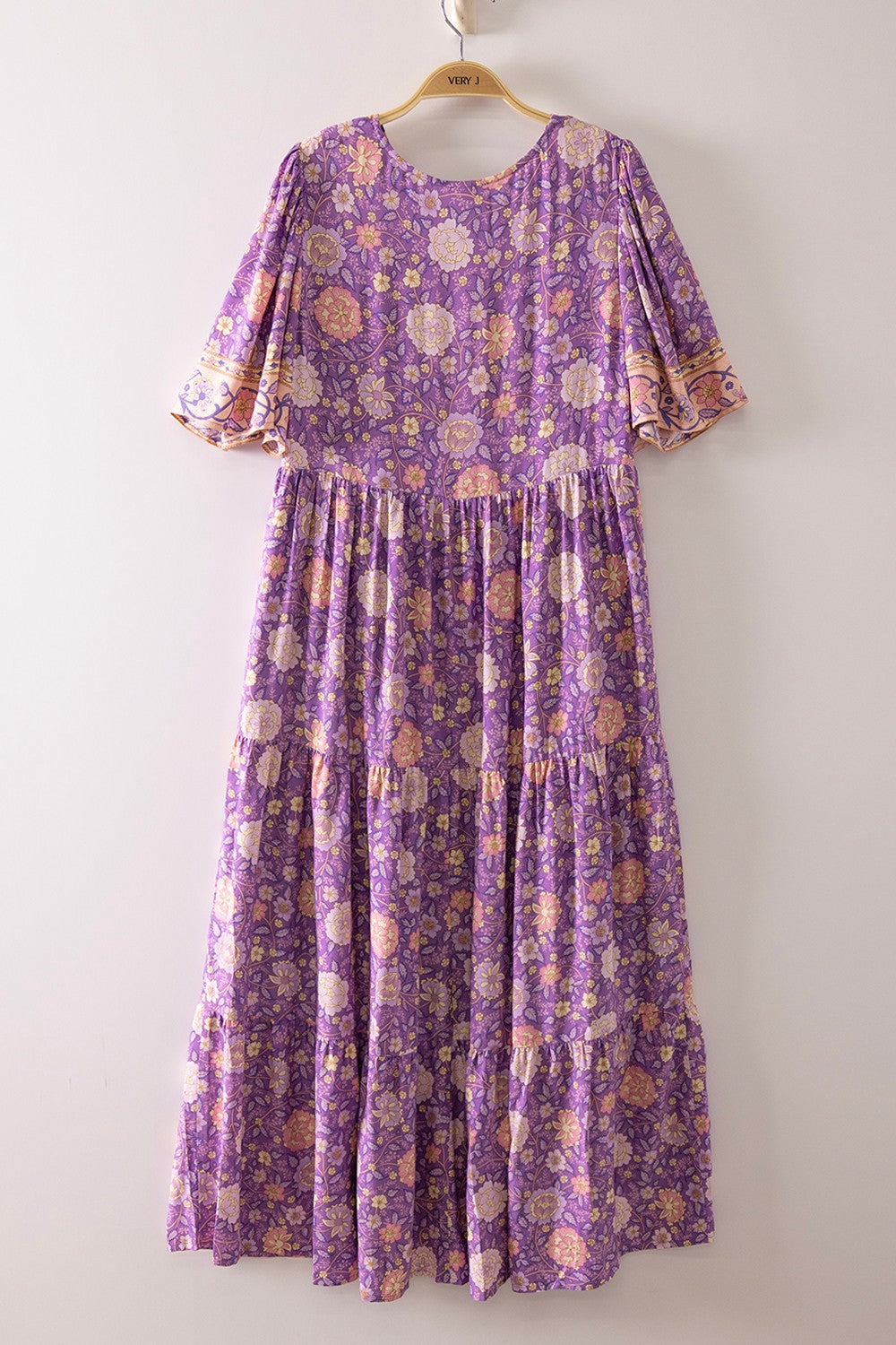 A short-sleeve tiered floral maxi dress in a lavender, coral, and gold mix featuring a deep V-neckline, button-down front closure, and decorative border print detailing.