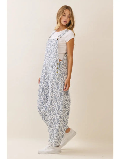 A women's sleeveless floral printed cropped overall jumpsuit in a blue and cream pattern featuring adjustable buckle shoulder straps, side button closures, utility slash pockets, and a relaxed wide-leg silhouette worn over a white short-sleeve tee.
